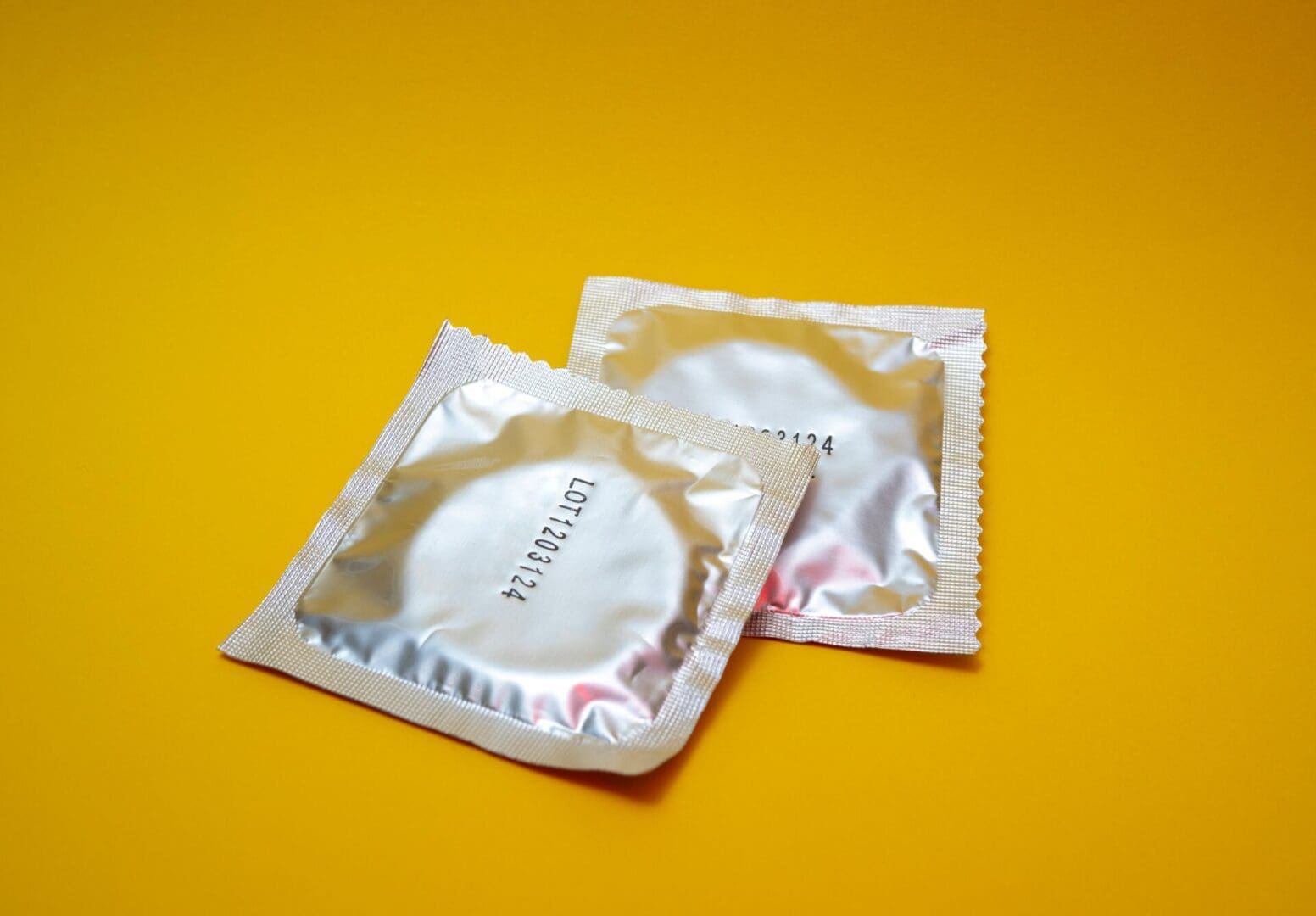 reproductive-health-supplies-coalition-RF5PjkWNYxU-unsplash (1) Image shows two condom wrappers in front of a yellow background