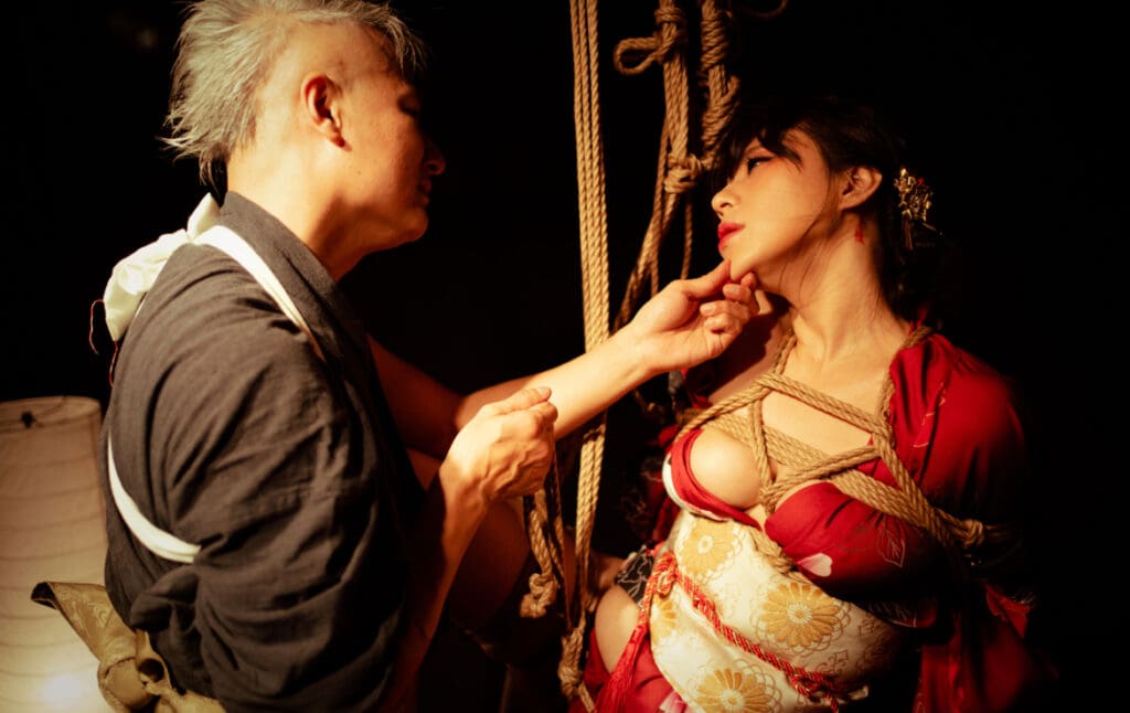 Shin holds chin of model bound by rope.